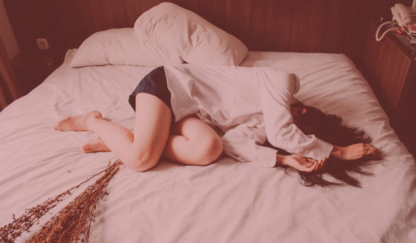 Frau liegt zusammengerollt quer auf einem Bett und hat sich unter die Bettdecke gezogen